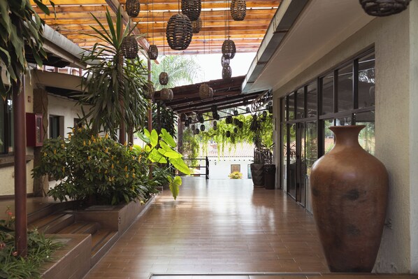 A covered walkway with wooden flooring, potted plants, and hanging wicker lamps. - Hotel Posada Virreyes (Tlaquepaque)
