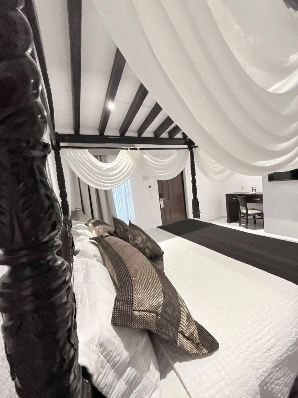 A bedroom with a four-poster bed, white bedding, and a view of a dining area.