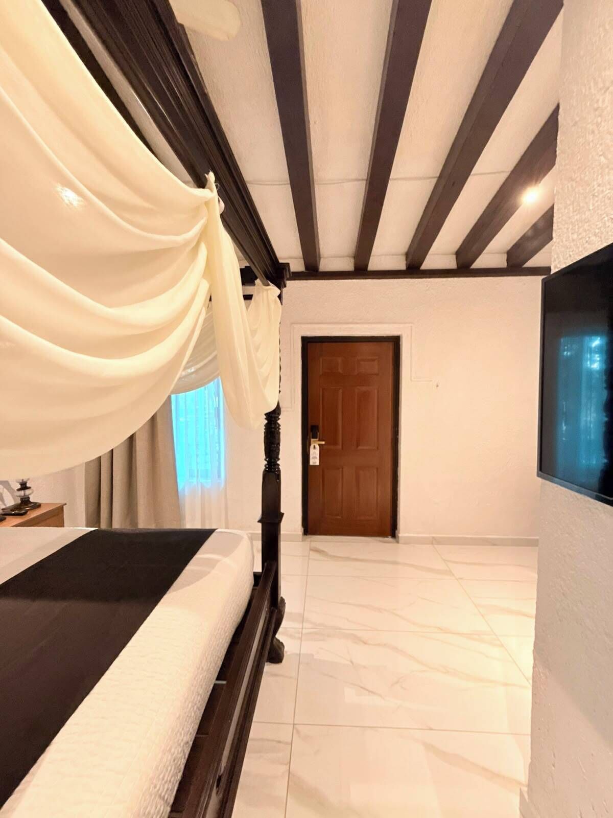 Une chambre d’hôtel avec un lit, une tête de lit en bois, une grande fenêtre avec des rideaux et une porte.