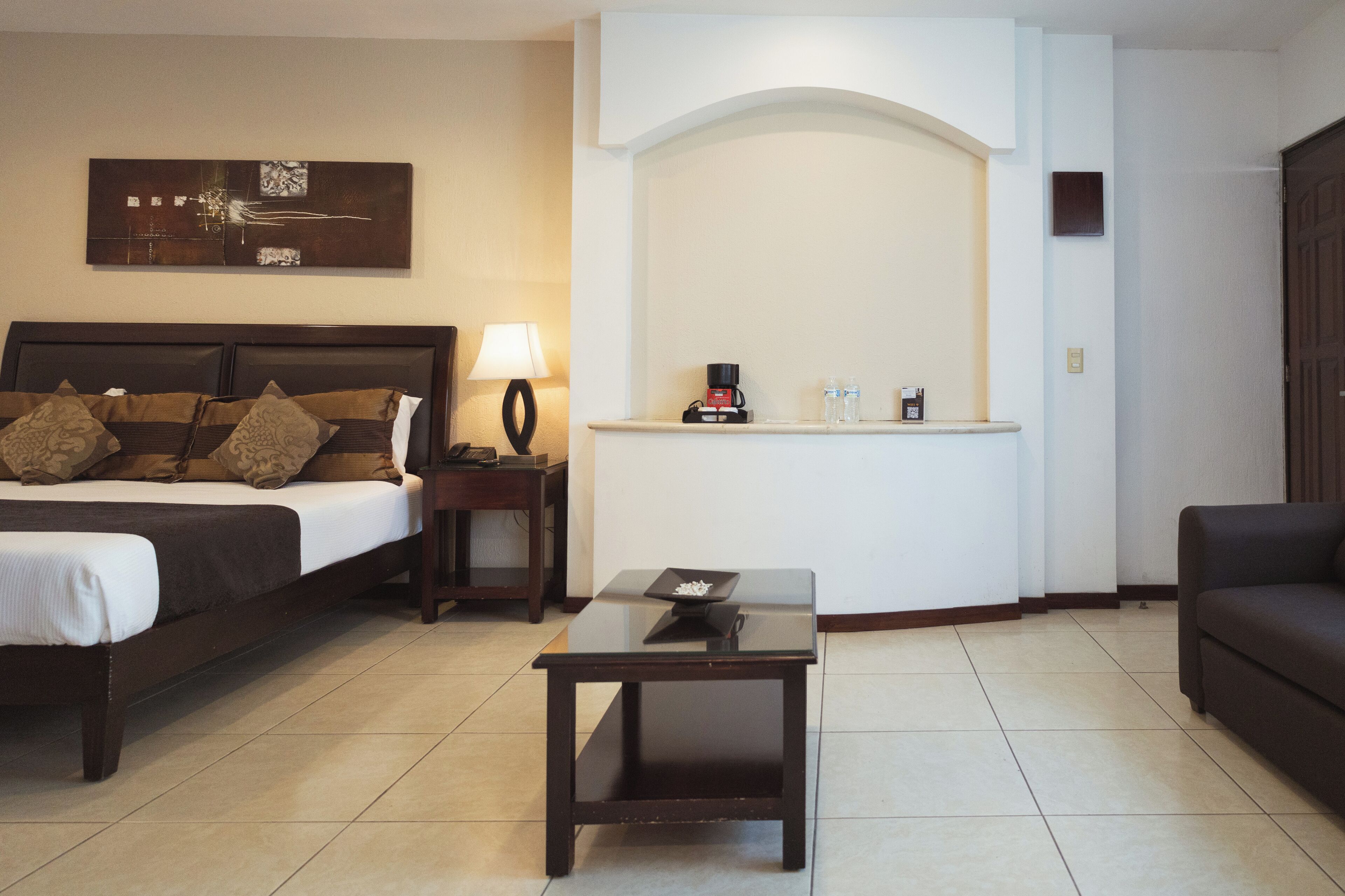 A hotel room with a bed, a small table, a lamp, a coffee maker, and a sofa.