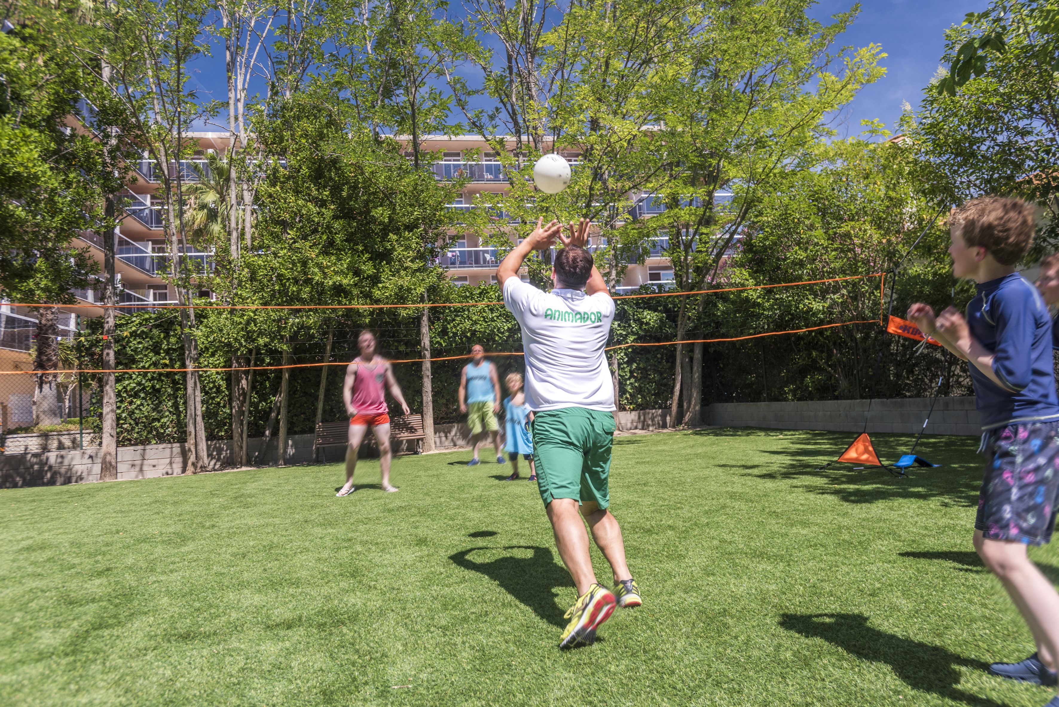 A person playing volleyball in a grassy area with trees and a building in the background.