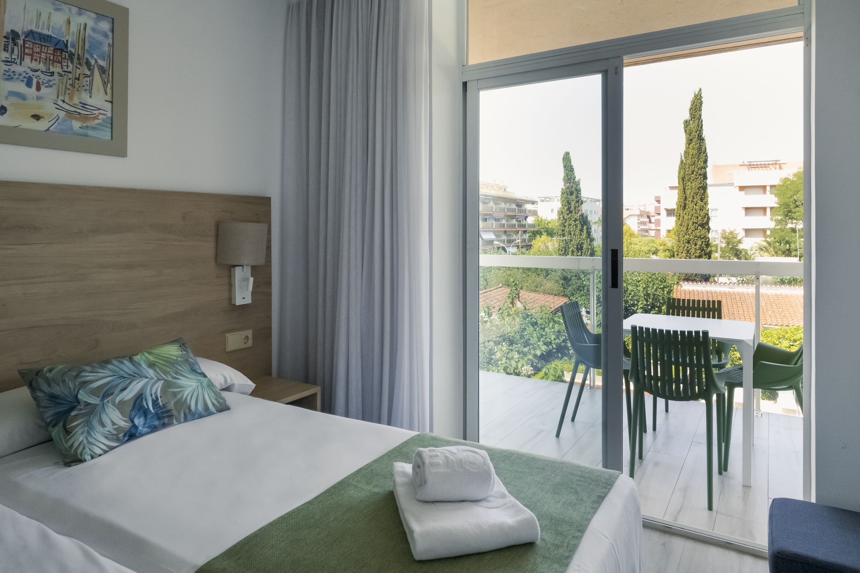 A hotel room with a bed, a green towel, a nightstand, a lamp, a painting, and a balcony with chairs and a table.