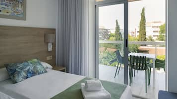 A hotel room with a bed, a green towel, a nightstand, a lamp, a painting, and a balcony with chairs and a table.