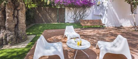 A patio with white chairs and a table, surrounded by a garden and a tree.