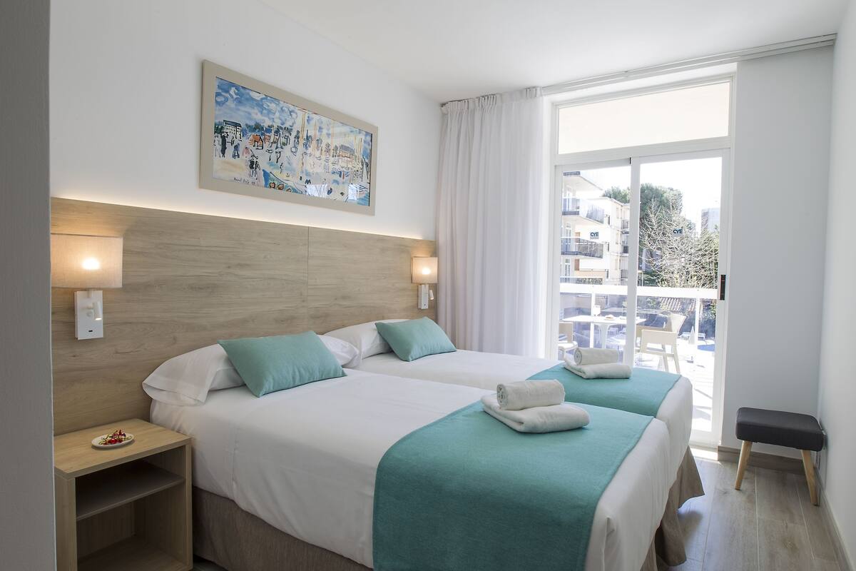 Quarto de hotel com duas camas, cabeceira de madeira, um quadro, varanda com vista para prédios e um banco.