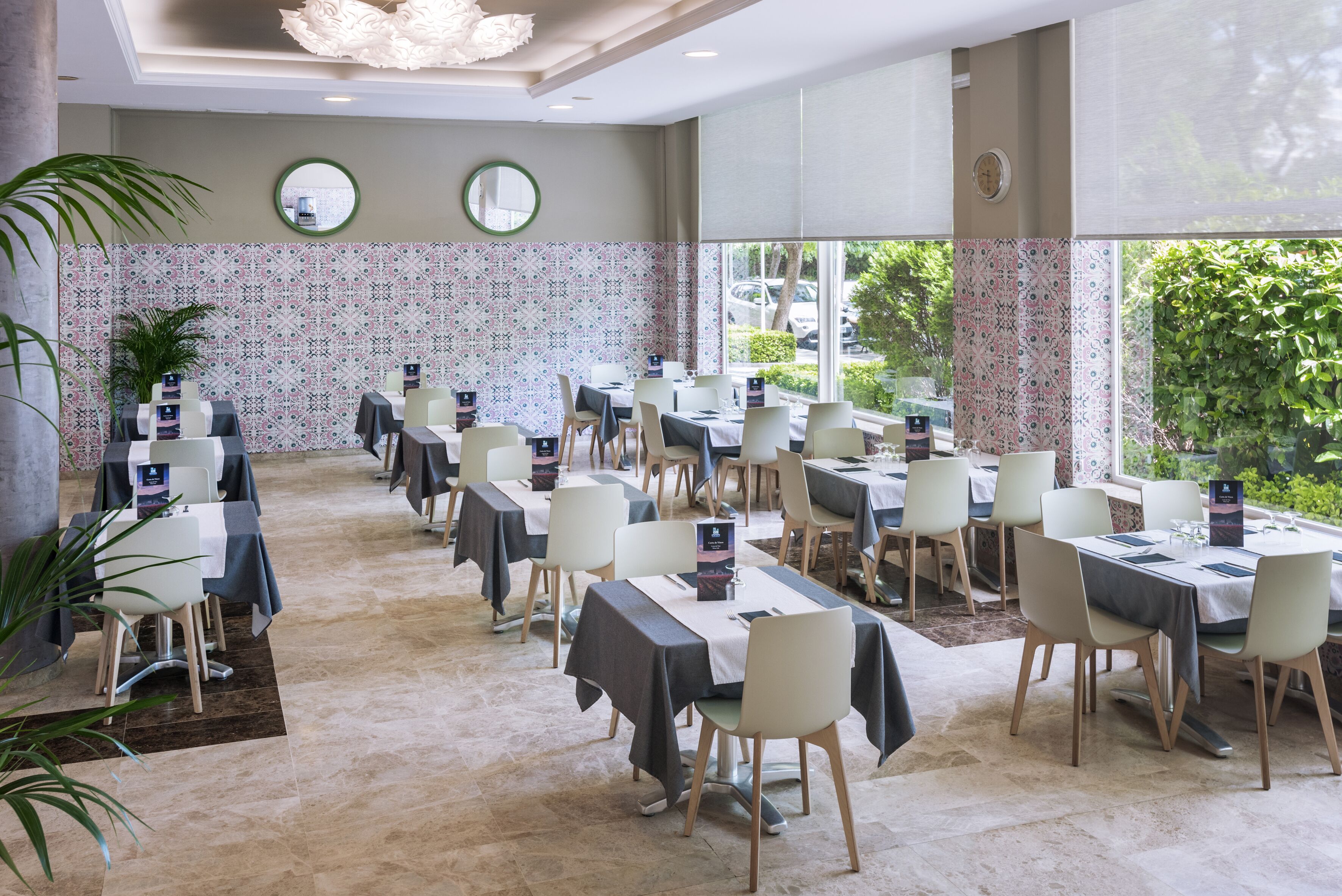 A dining area with multiple tables and chairs, large windows with a view of greenery, and decorative wall tiles.