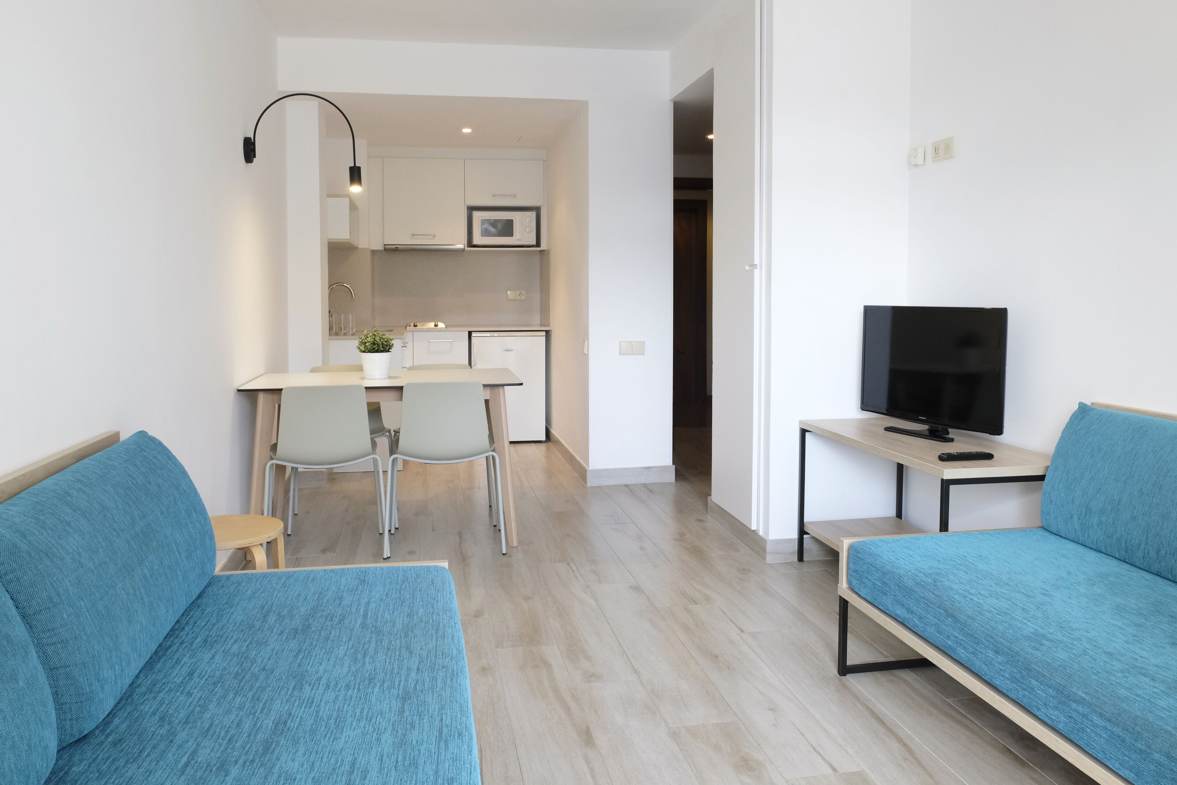 A modern living room with teal sofas, a TV on a wooden table, and a kitchen in the background.