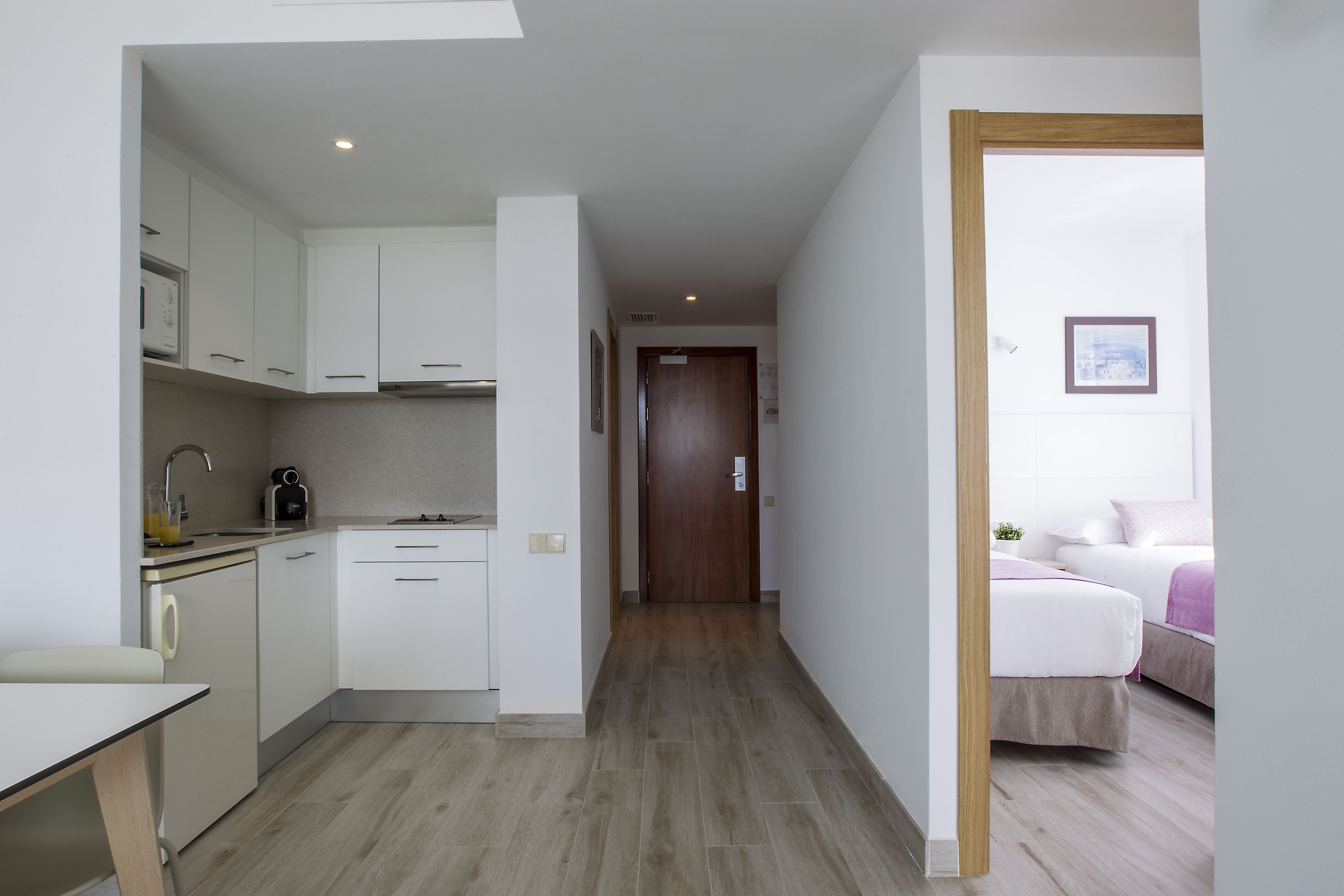 A modern kitchen with white cabinets, a sink, and a coffee machine. A hallway leads to a bedroom with a bed and a window.