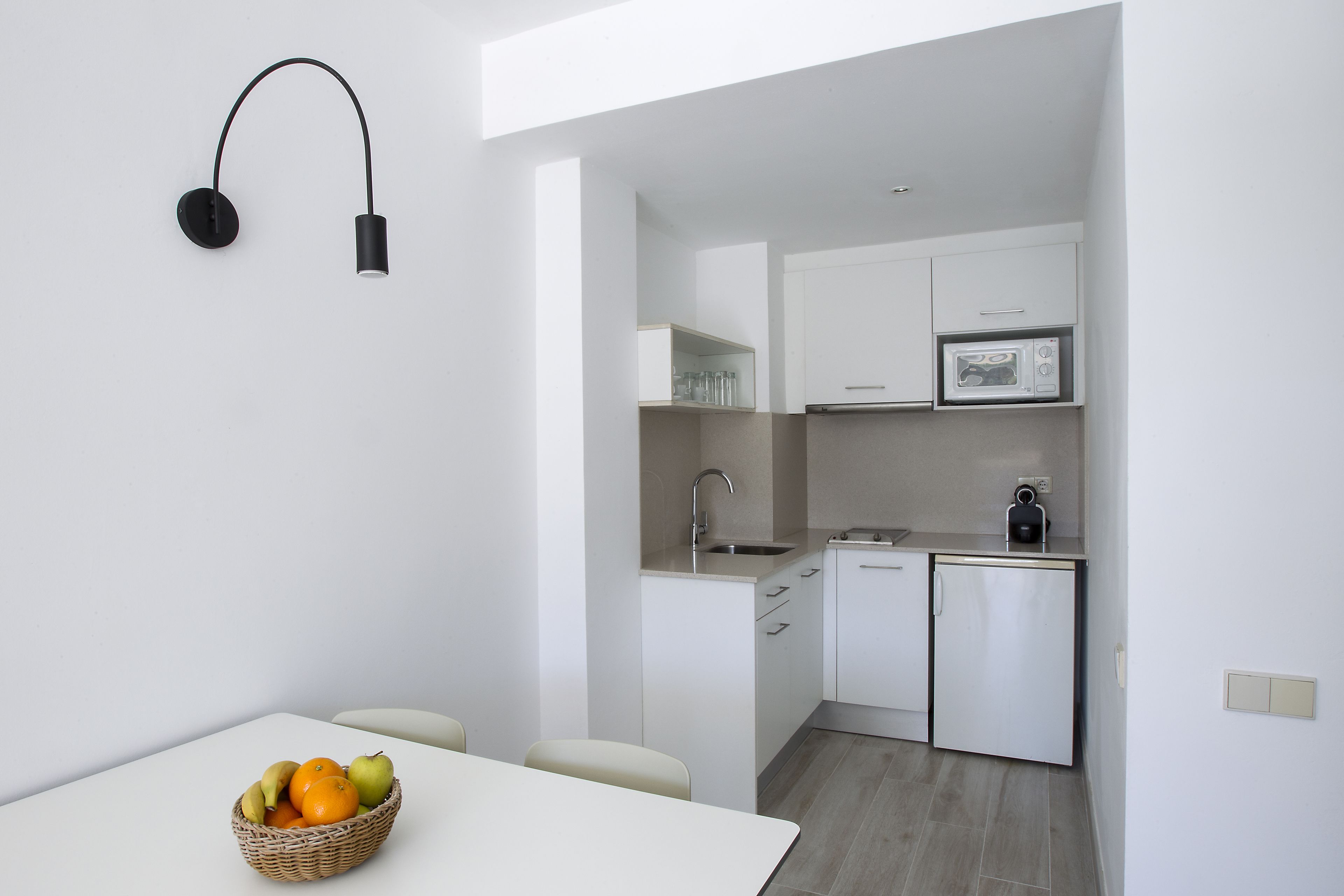A compact kitchen with white cabinets, a microwave, and a small refrigerator. There is a wicker basket with fruits on the table.