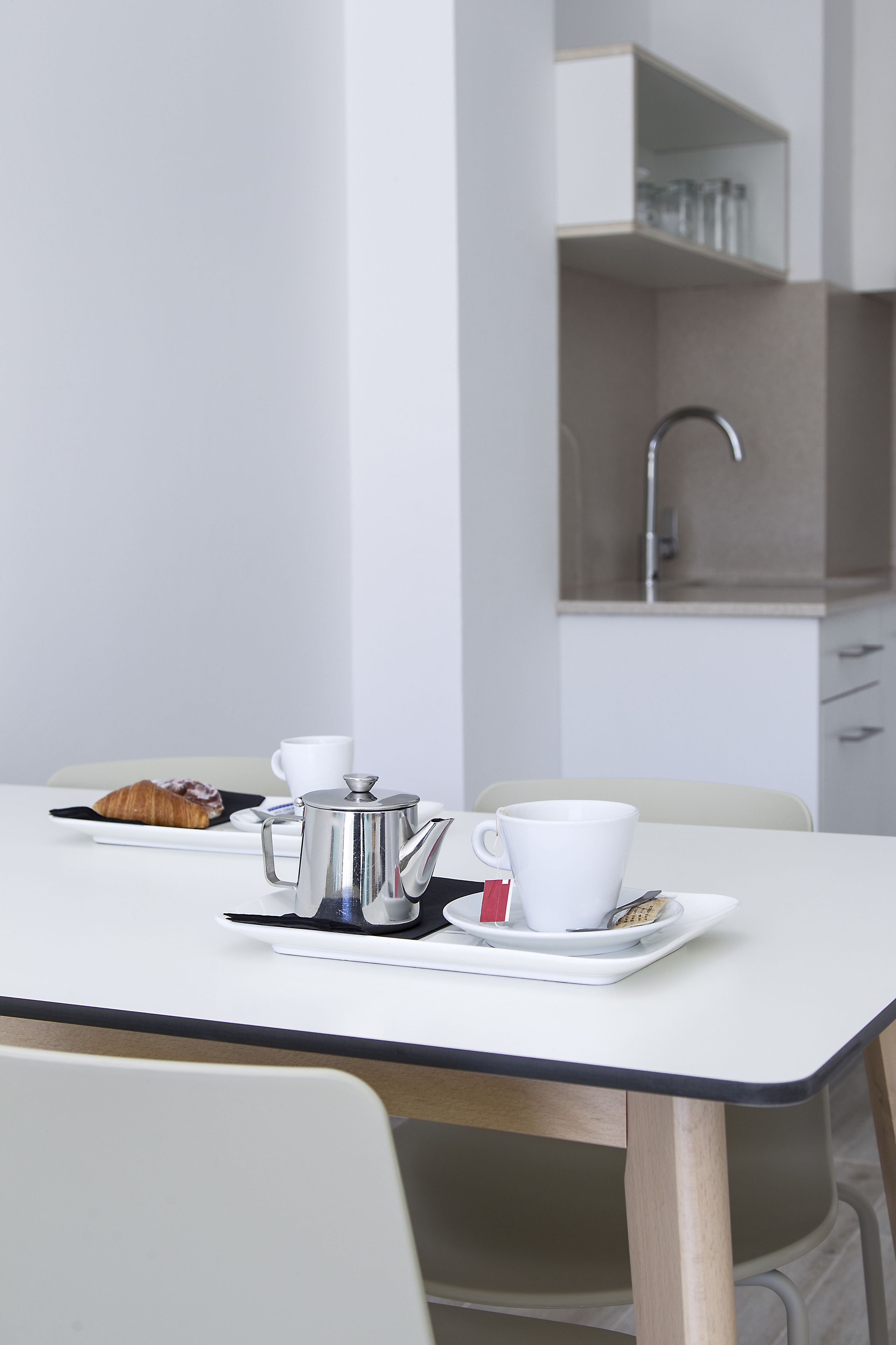 Una cocina moderna con encimera blanca, cafetera de acero inoxidable, taza blanca y un croissant sobre un plato negro.