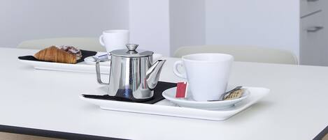 A modern kitchen with a white countertop, a stainless steel coffee pot, a white cup, and a croissant on a black plate.