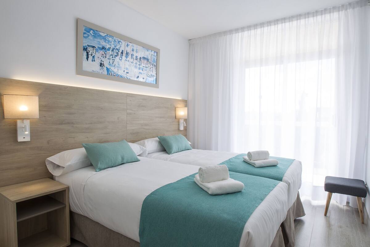 Quarto de hotel com duas camas, cabeceira de madeira, pintura na parede e um banco junto à janela.