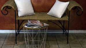 A wicker chair with white cushions and a round metal table with magazines. - Hôtel Le Petit Château (Cannes)