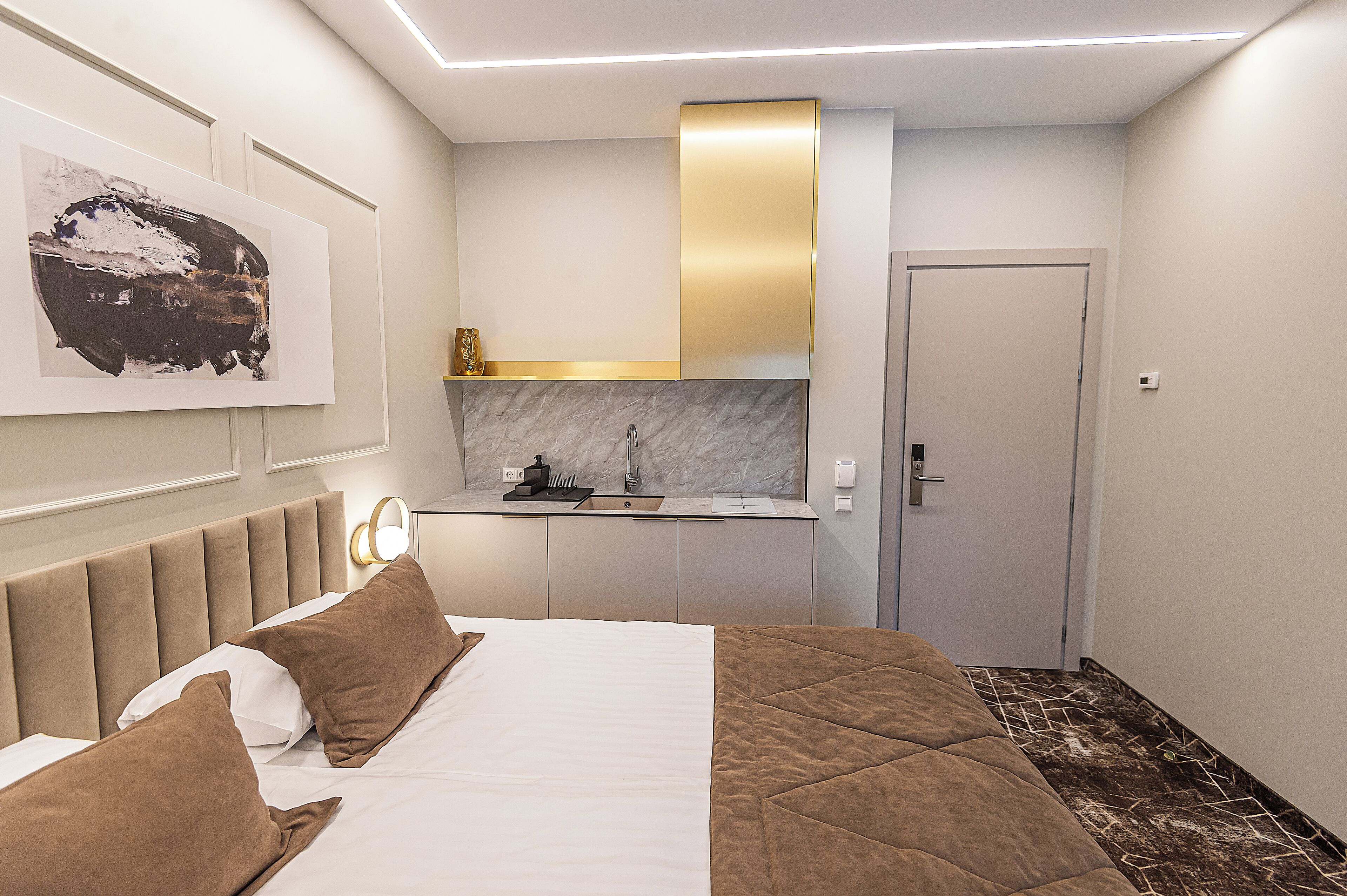 Una habitación de hotel moderna con una cama, una mesita de noche, un espejo, un cuadro, una kitchenette y una puerta.