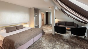 Habitación de hotel moderna con una cama grande, zona de estar con sofá y sillones, y vistas al exterior.