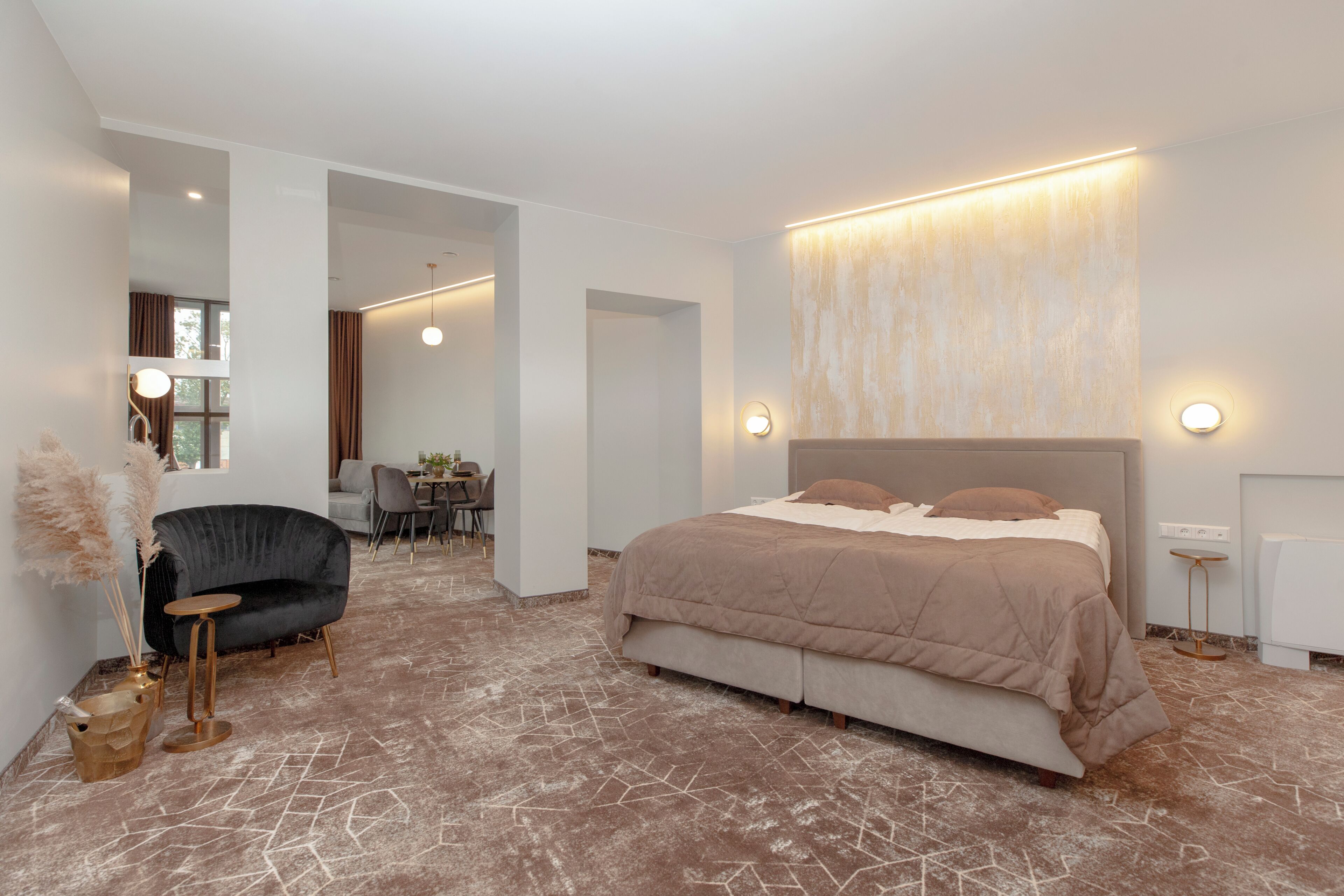 A modern bedroom with a large bed, a curved sofa, a small table, and a dining area in the background.