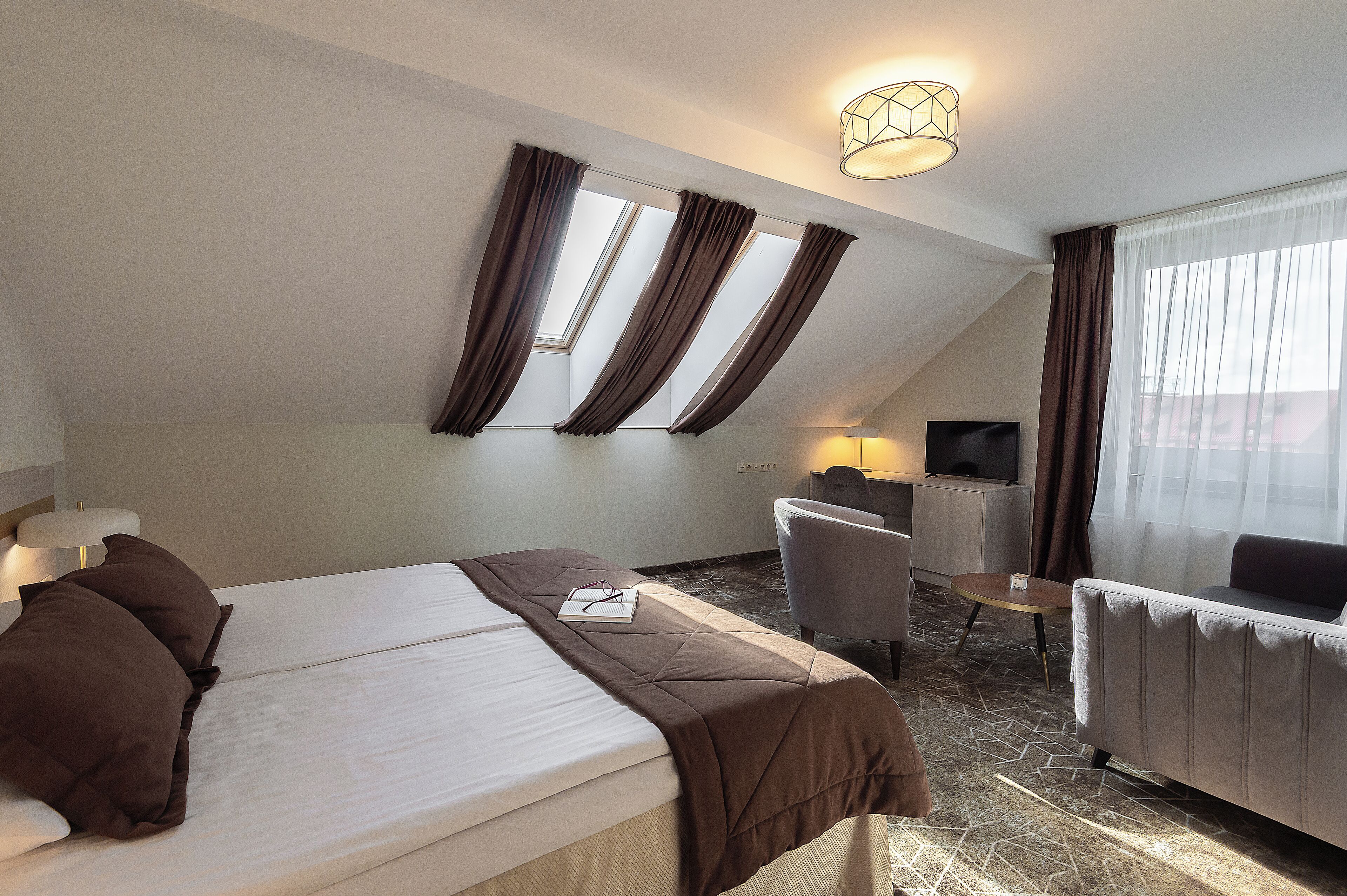 Habitación de hotel con cama, escritorio, sillón y sofá. Cuenta con ventanas con cortinas, una lámpara en el techo y un televisor sobre el escritorio.