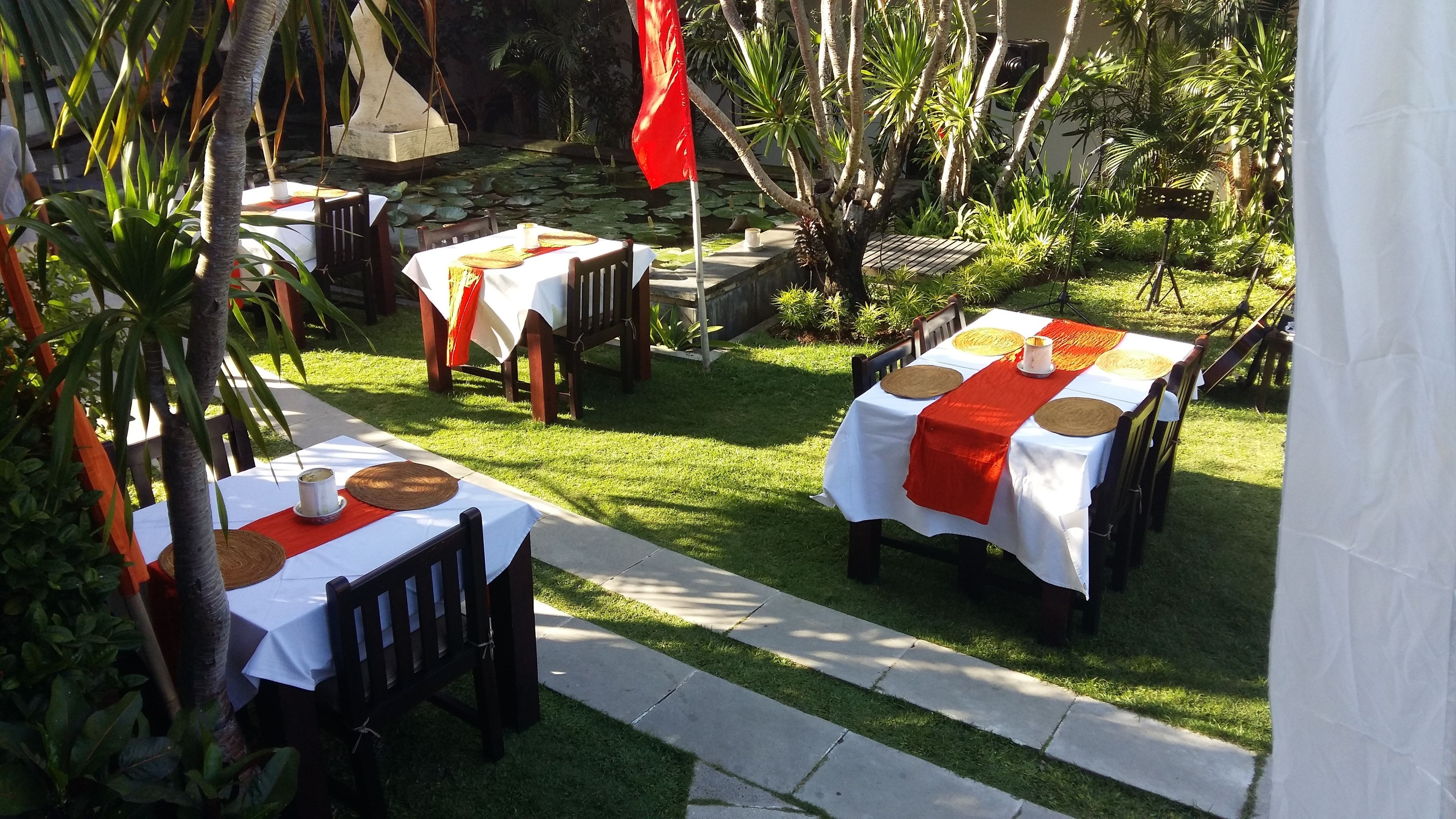An outdoor dining area with tables set up on a grassy lawn, surrounded by lush greenery and a stone structure.