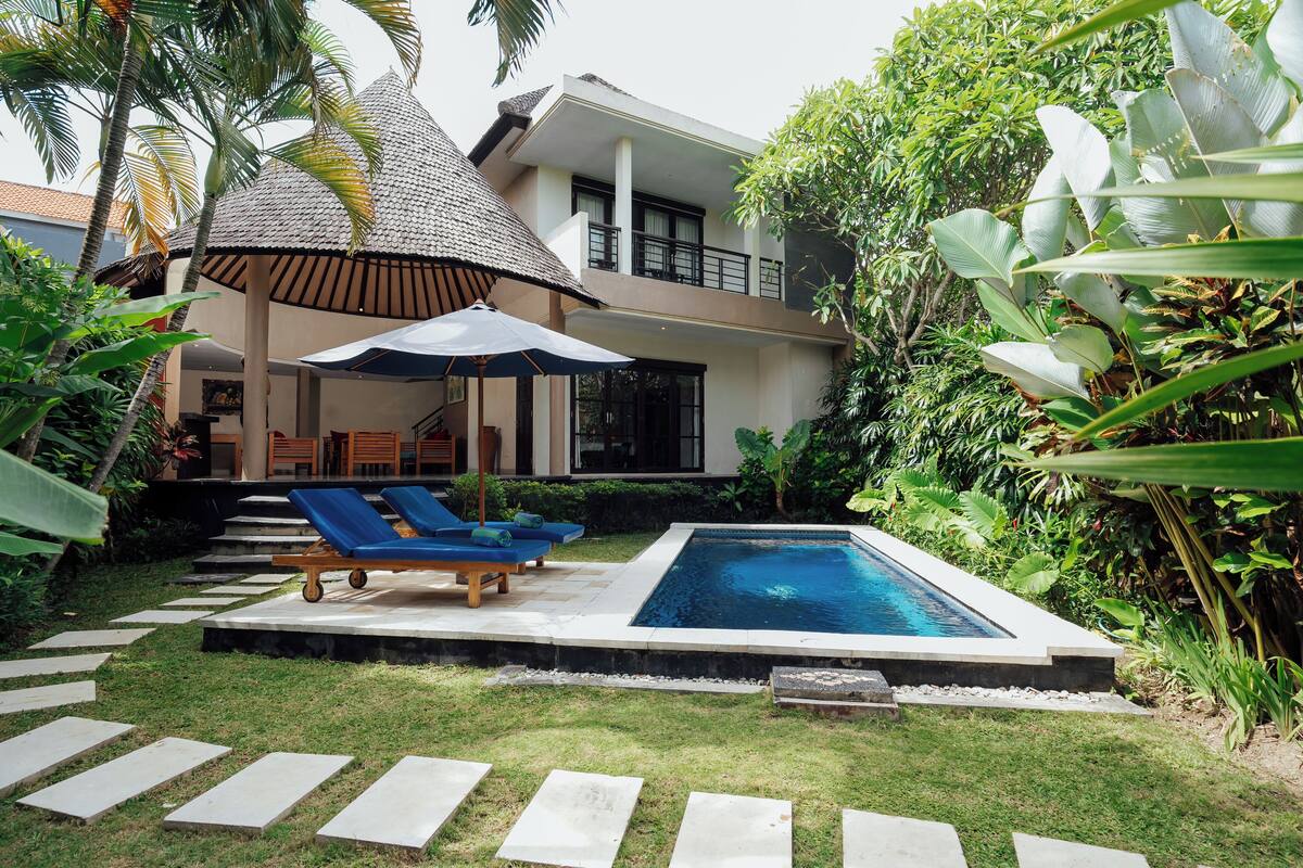 Ein modernes Haus mit Swimmingpool, Liegestühlen und einem strohgedeckten Gebäude.