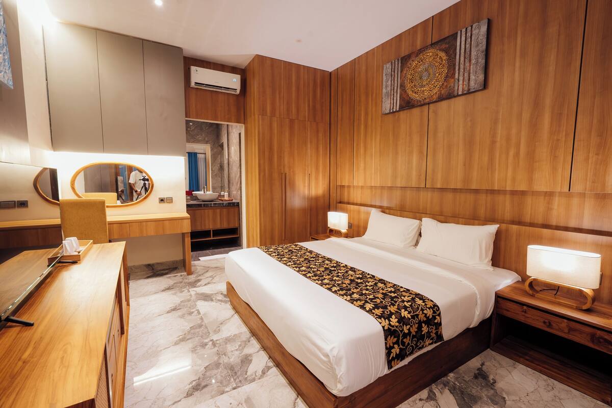 Ein modernes Hotelzimmer mit einem Holzkopfteil, einem großen Bett mit weißen Leinentüchern, einem Schreibtisch mit Spiegel und einem Nachttisch mit Lampe.