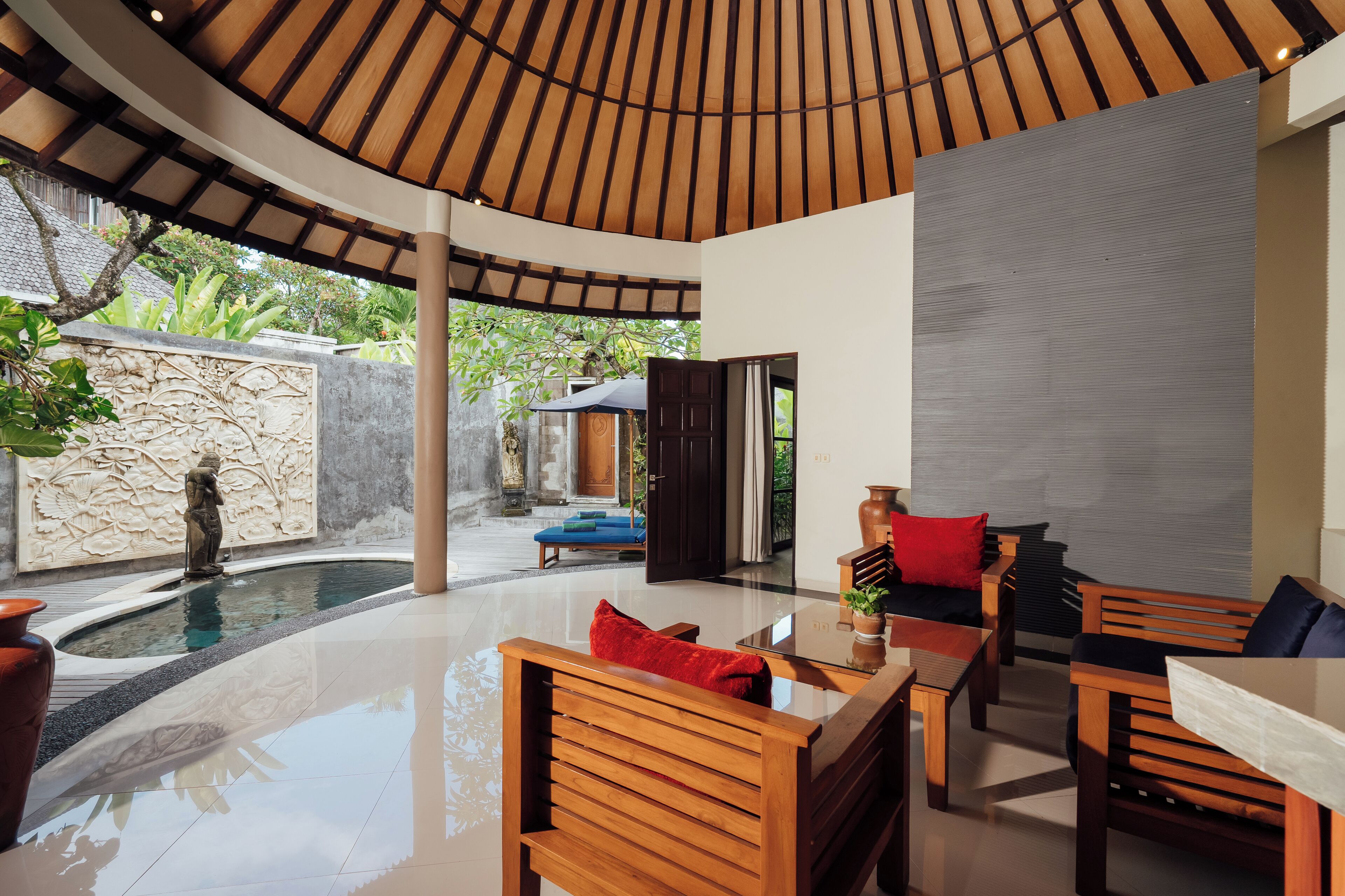 Un espacio interior moderno con techo de madera curvada, pared de piedra con relieve tallado, una pequeña piscina y muebles de madera con cojines.