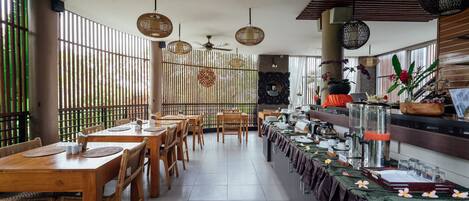 Un restaurante con mesas y sillas de madera, mostrador de buffet, ventiladores de techo e iluminación decorativa colgante.