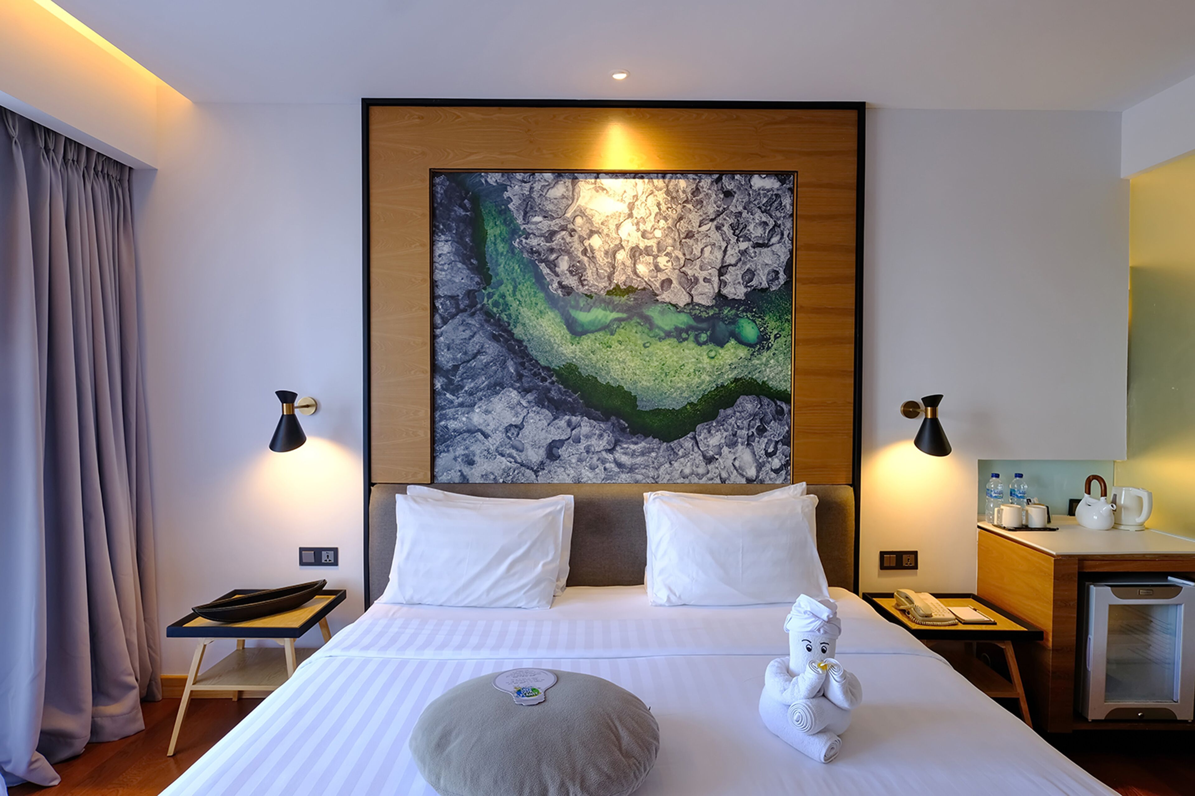 A hotel room with a large bed, two bedside lamps, a painting on the wall, a small desk with a kettle, and a minibar.