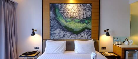 A hotel room with a large bed, two bedside lamps, a painting on the wall, a small desk with a kettle, and a minibar.