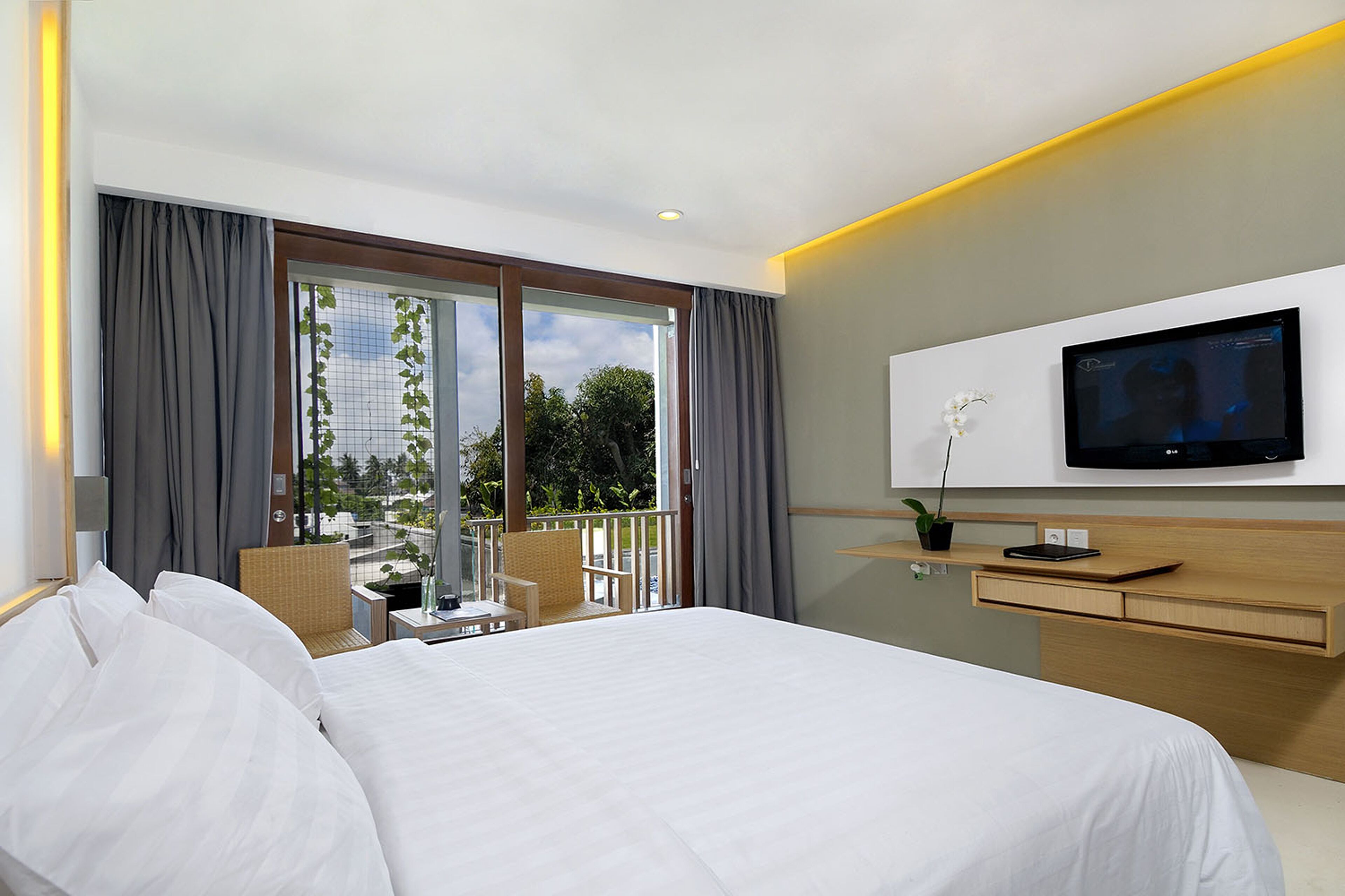 A hotel room with a large bed, a television mounted on the wall, a small desk with a plant, and a balcony with a chair and a table.