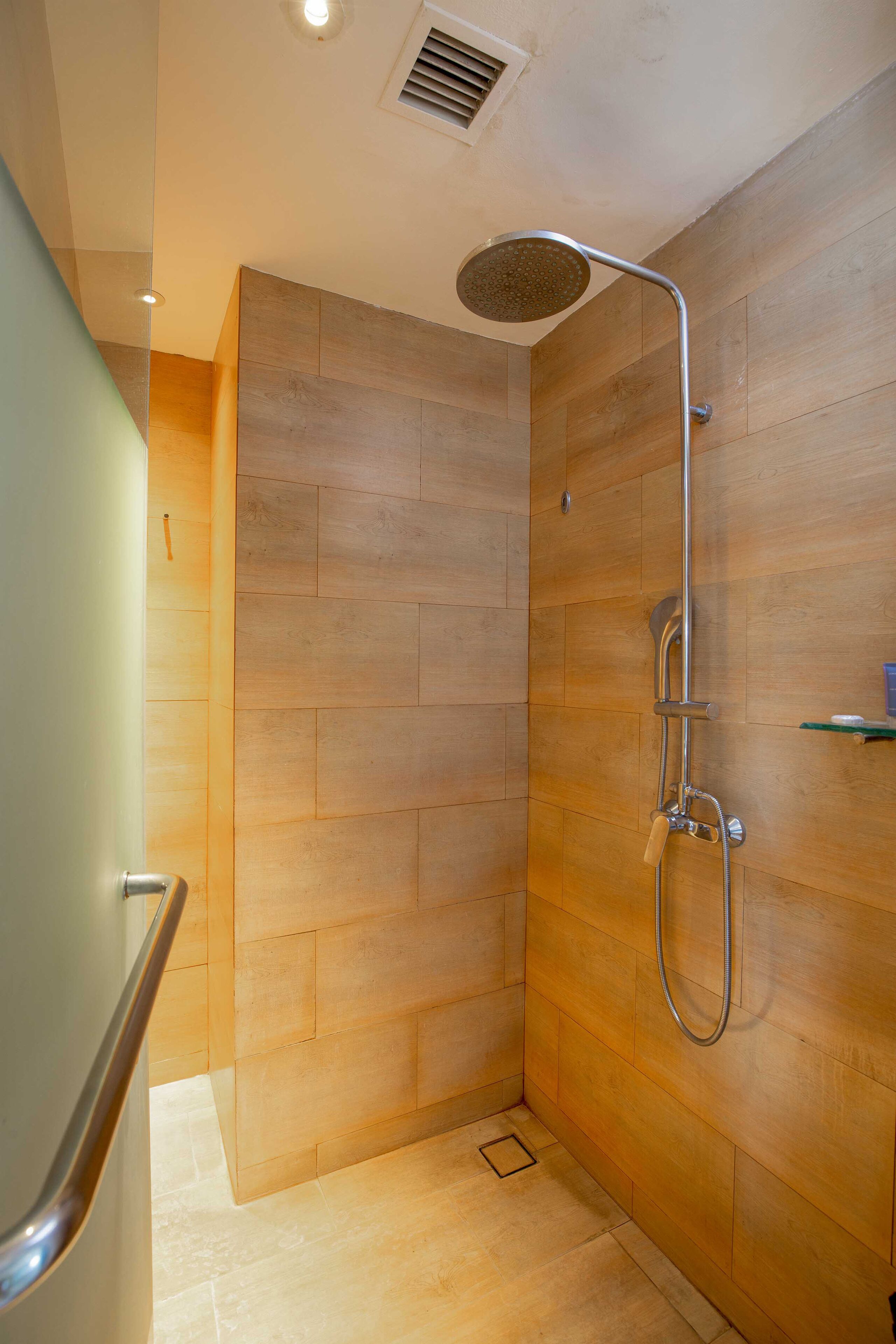 A modern shower with a handheld showerhead and a wall-mounted faucet, wooden paneling, and a small shelf with toiletries.