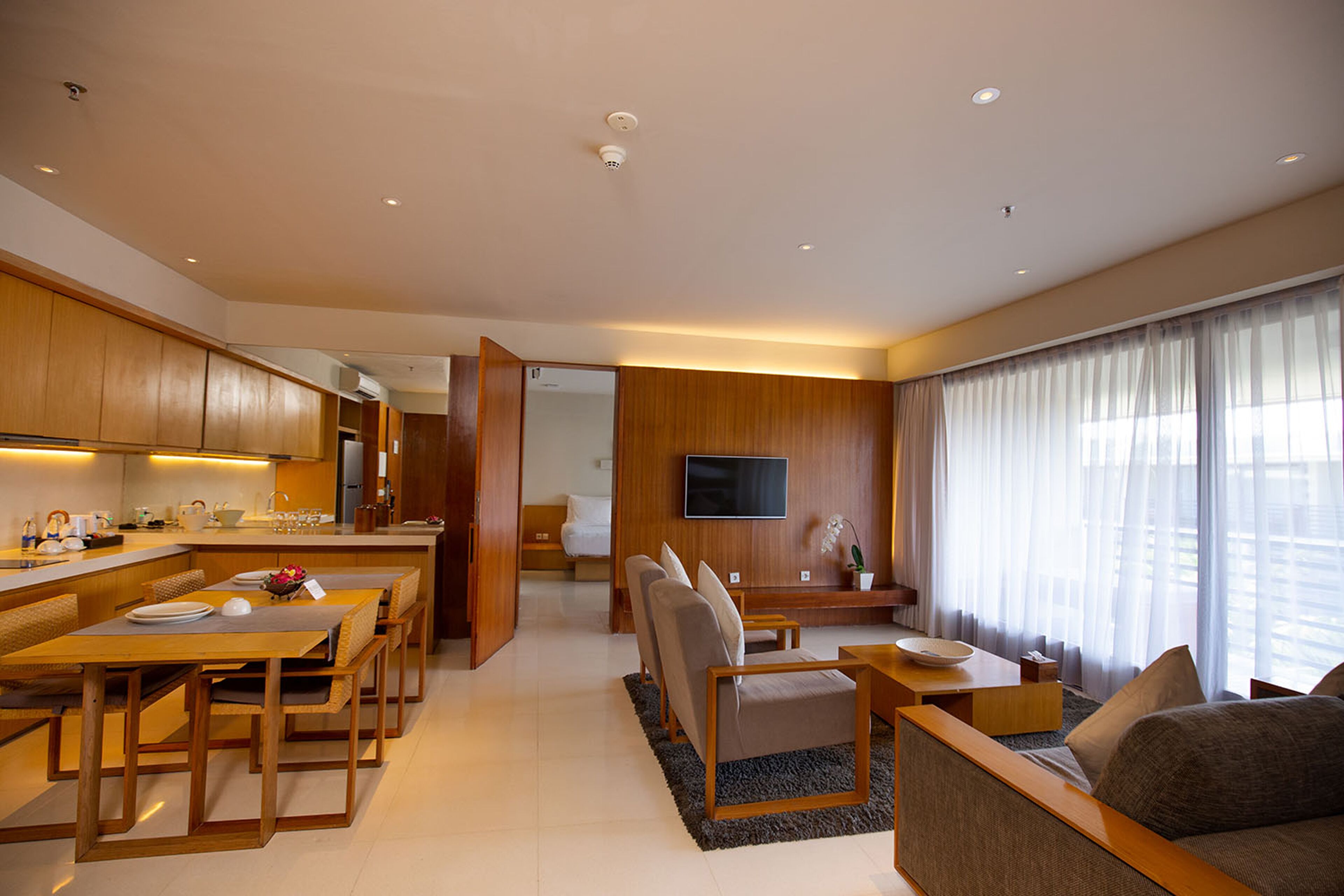 A modern hotel room with a dining area, a living room with a sofa and armchairs, a TV mounted on the wall, and a large window with sheer curtains.