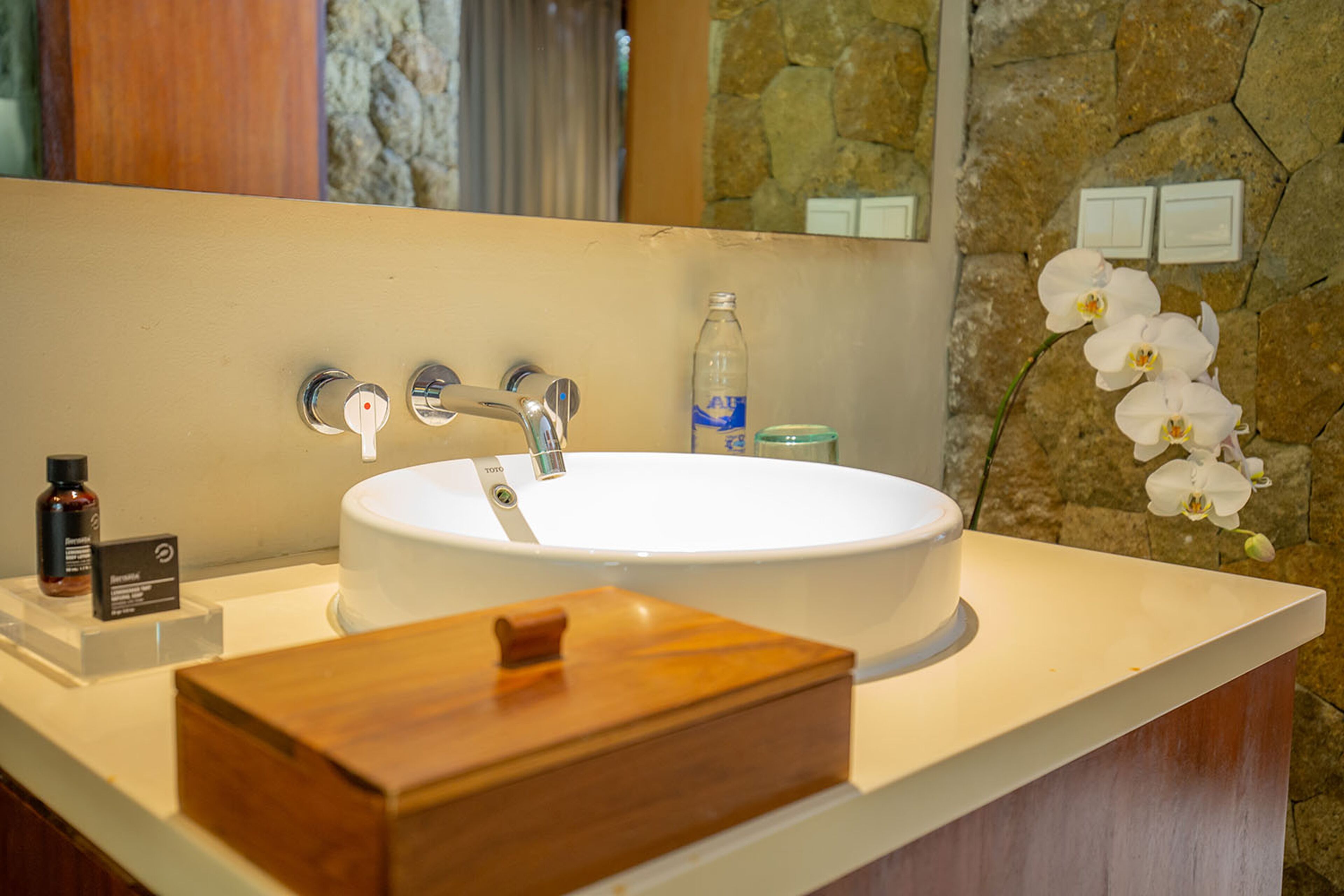 A bathroom with a large oval sink, a wooden box, a bottle of hand soap, and a potted orchid.