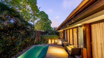 A wooden deck with a swimming pool, a lounge area with cushions, and a building with large windows.