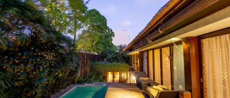 Una terraza de madera con piscina, un espacio de descanso con cojines y un edificio con ventanas amplias.
