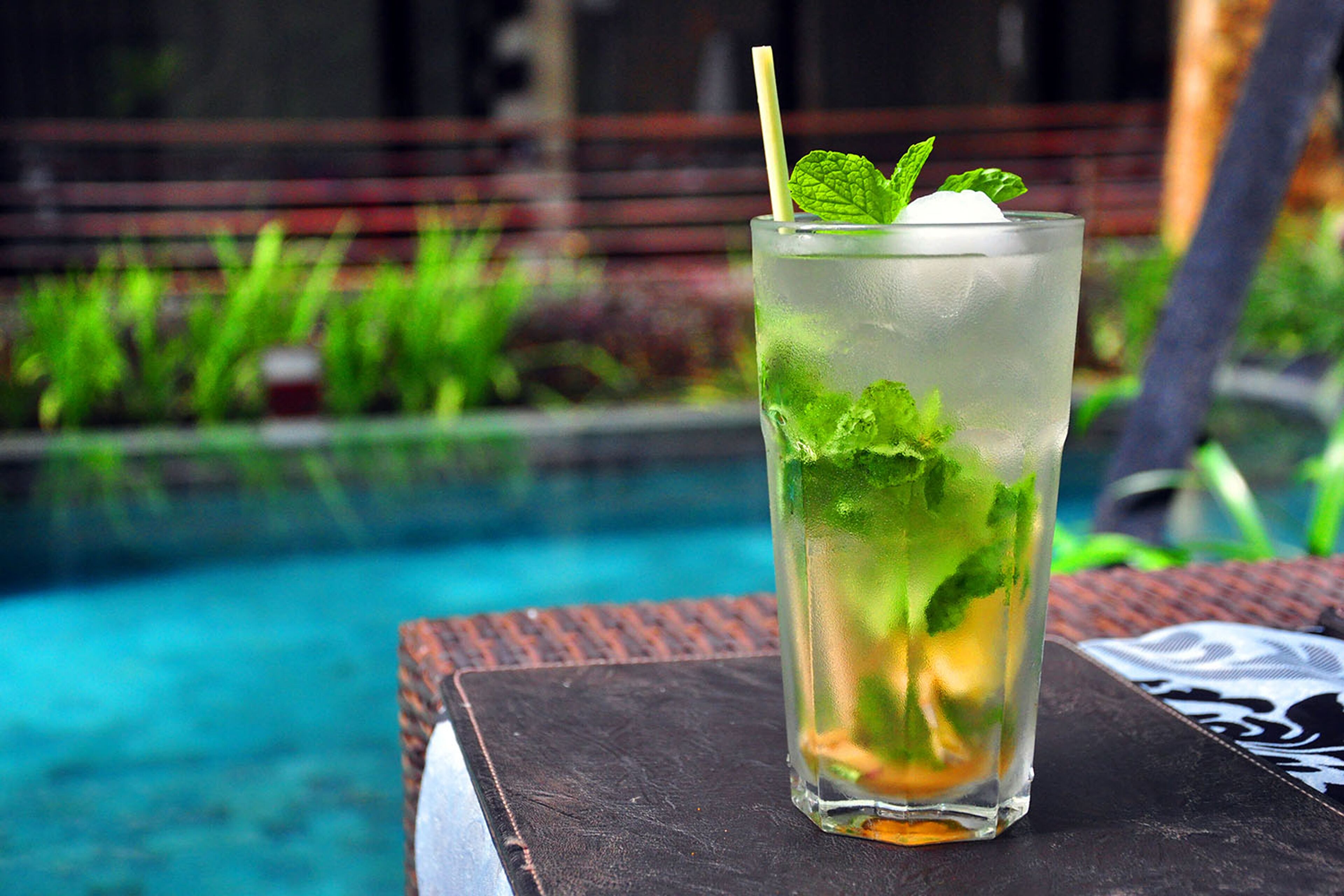 A tall glass with ice, mint leaves, and a straw, placed on a dark surface near a pool.