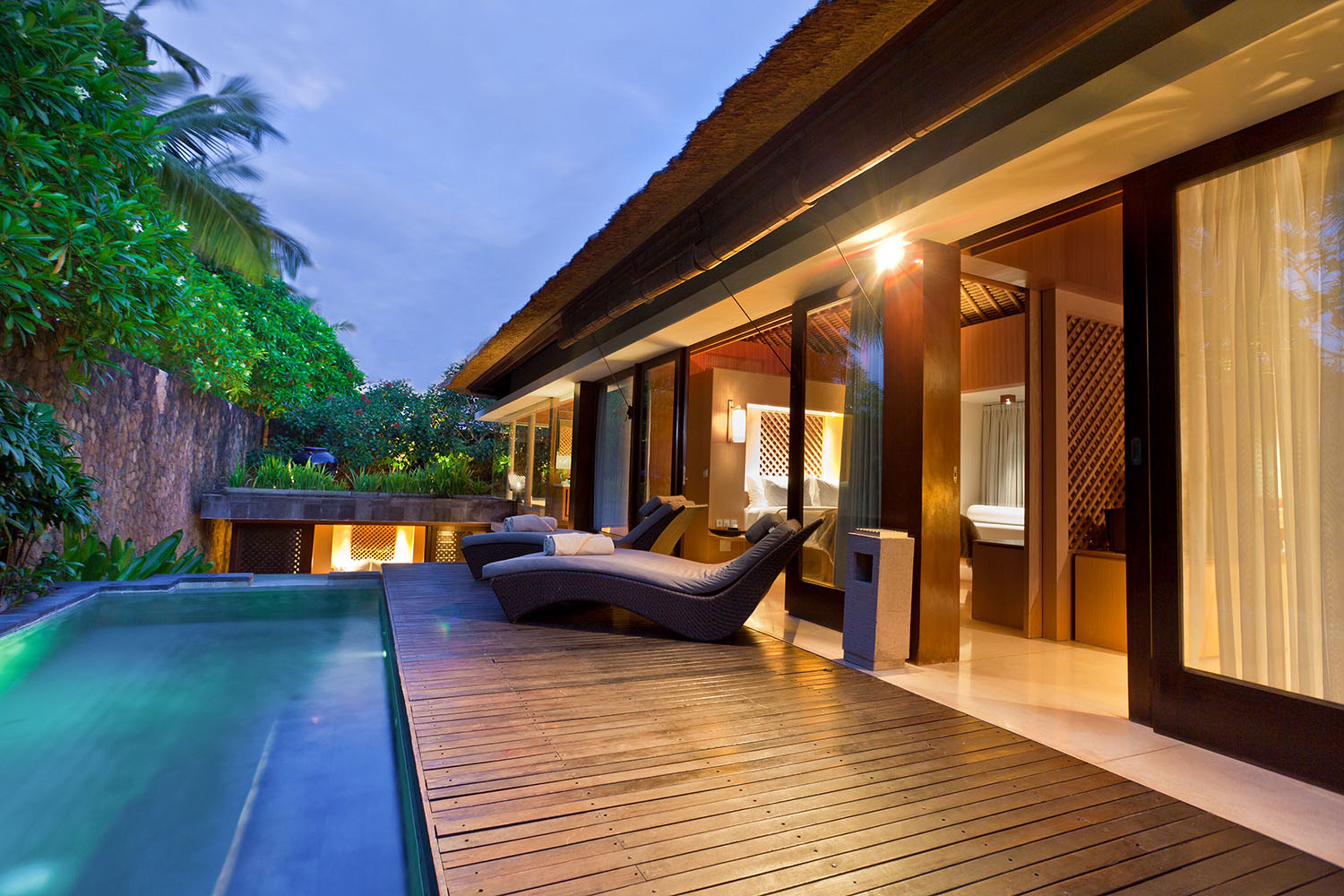 A modern outdoor pool area with lounge chairs, a wooden deck, and a well-lit interior space.