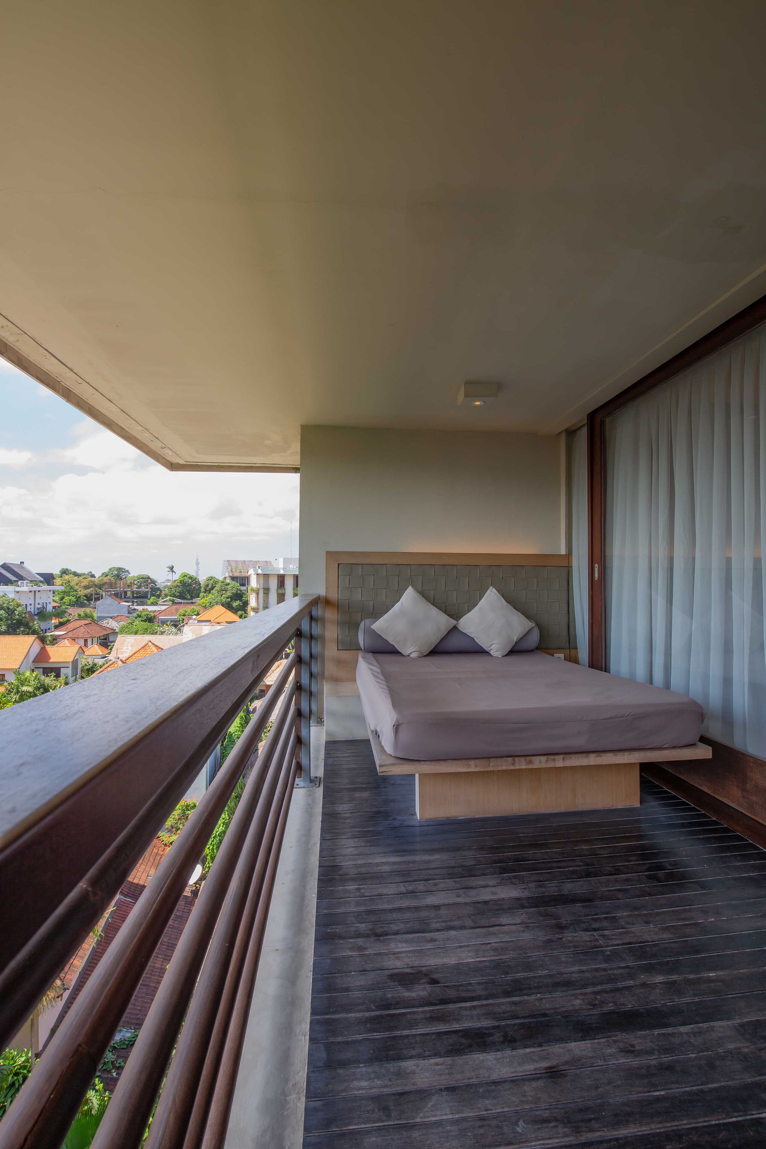 A balcony with a bed, wooden floor, and a view of buildings and trees.