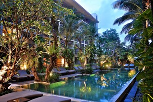 Uma piscina infinita com espreguiçadeiras e plantas tropicais, rodeada por um edifício com varandas.