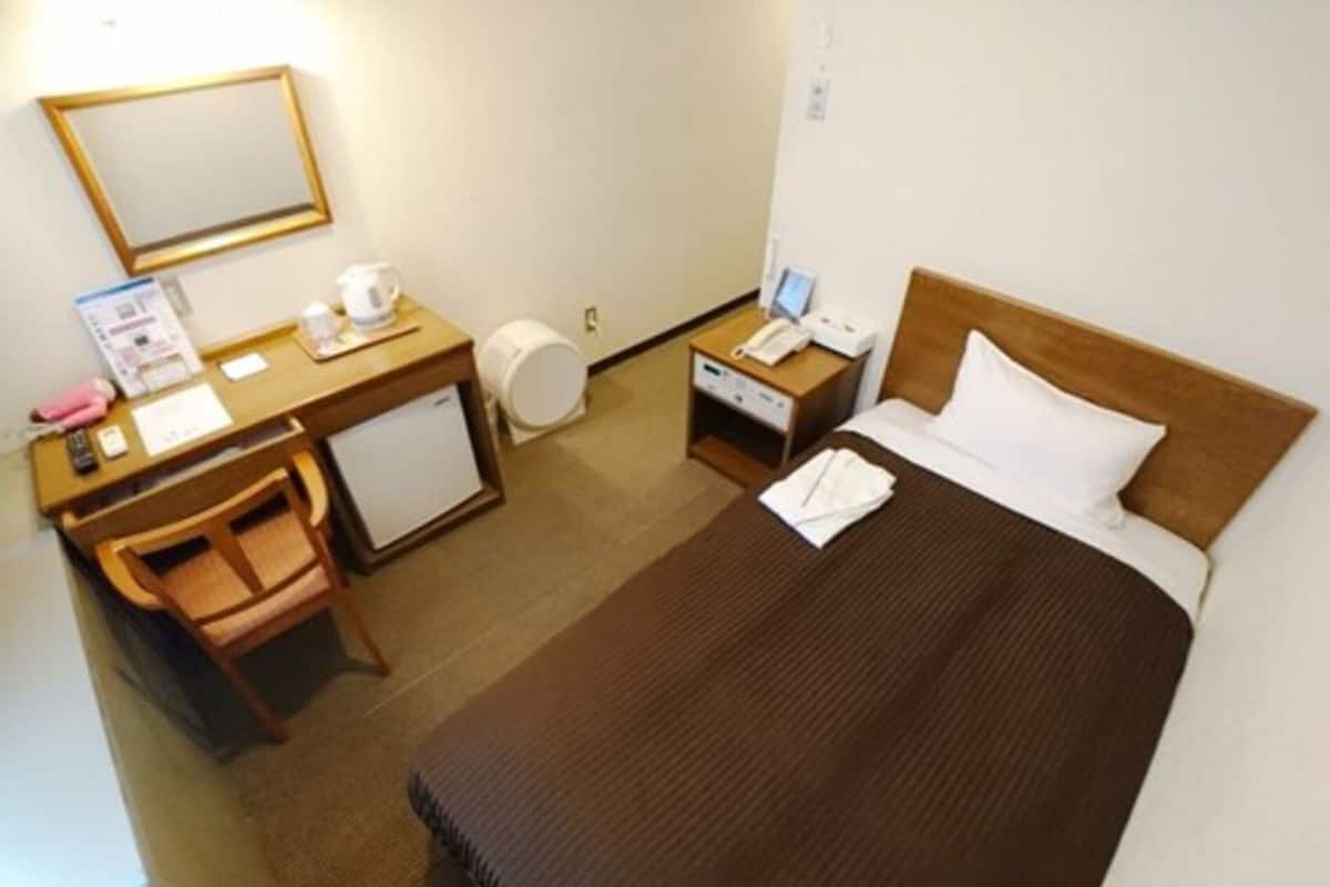 A hotel room with a bed, a desk, a chair, a small refrigerator, a bedside table with a lamp, and a mirror on the wall.