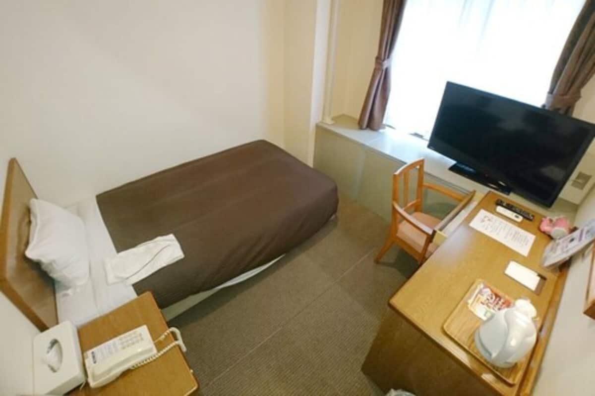 A small hotel room with a single bed, a desk with a television, a chair, and a small table with a cup and papers.