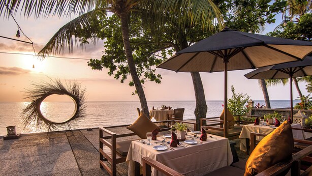 Un espace de restauration en plein air avec des tables et des chaises, un grand parasol et une vue sur l’océan au coucher du soleil.