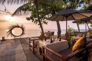 An outdoor dining area with tables and chairs, a large umbrella, and a view of the ocean at sunset.