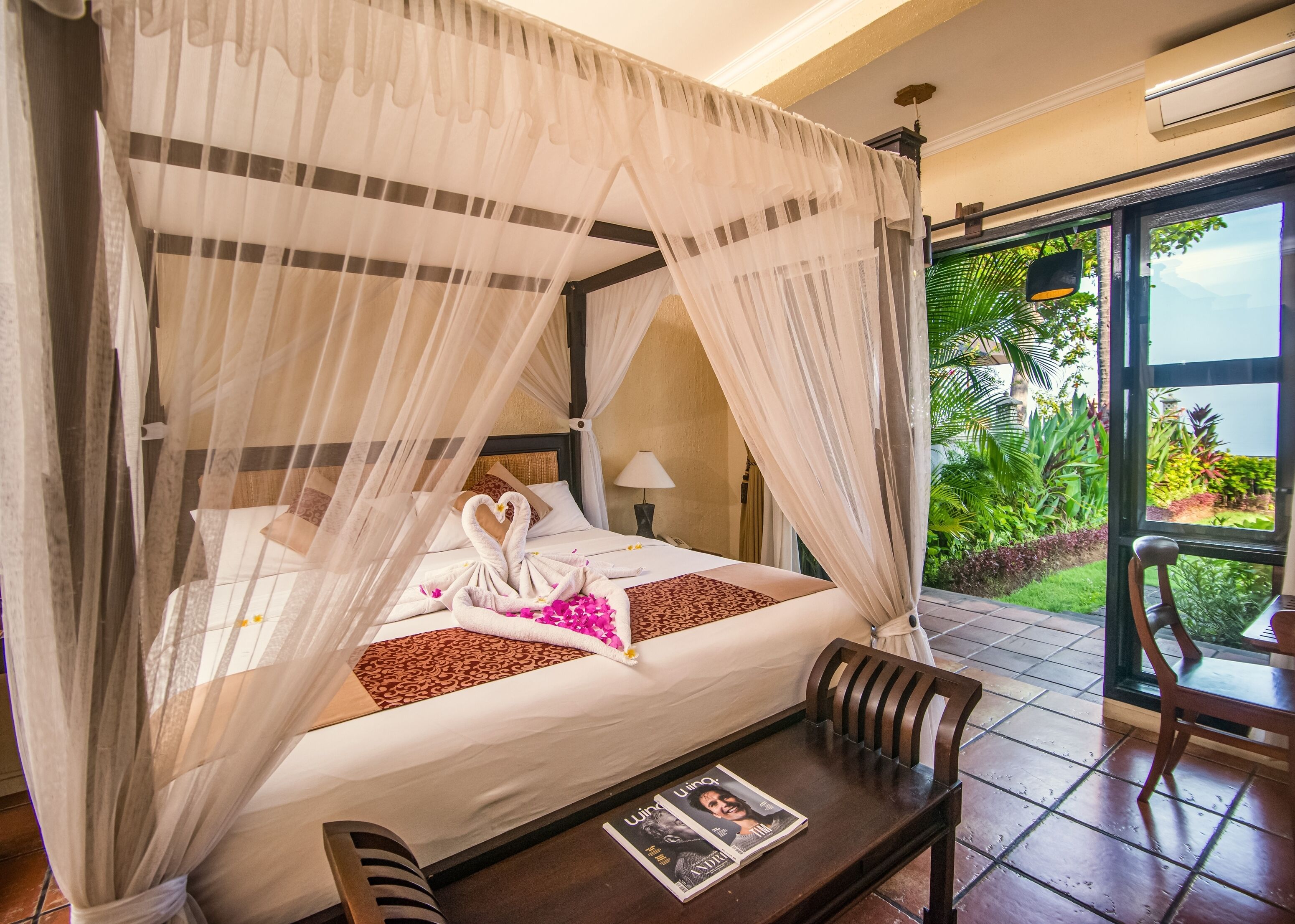 A four-poster bed with a canopy, a heart-shaped towel arrangement, a magazine on the table, and a view of lush greenery outside.
