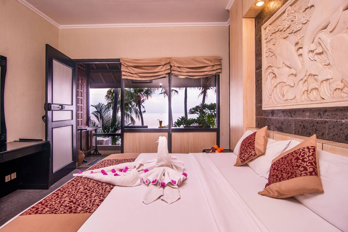 Habitación de hotel con cama, almohadas, televisor, ventana y vista a palmeras, además de un relieve decorativo en la pared.
