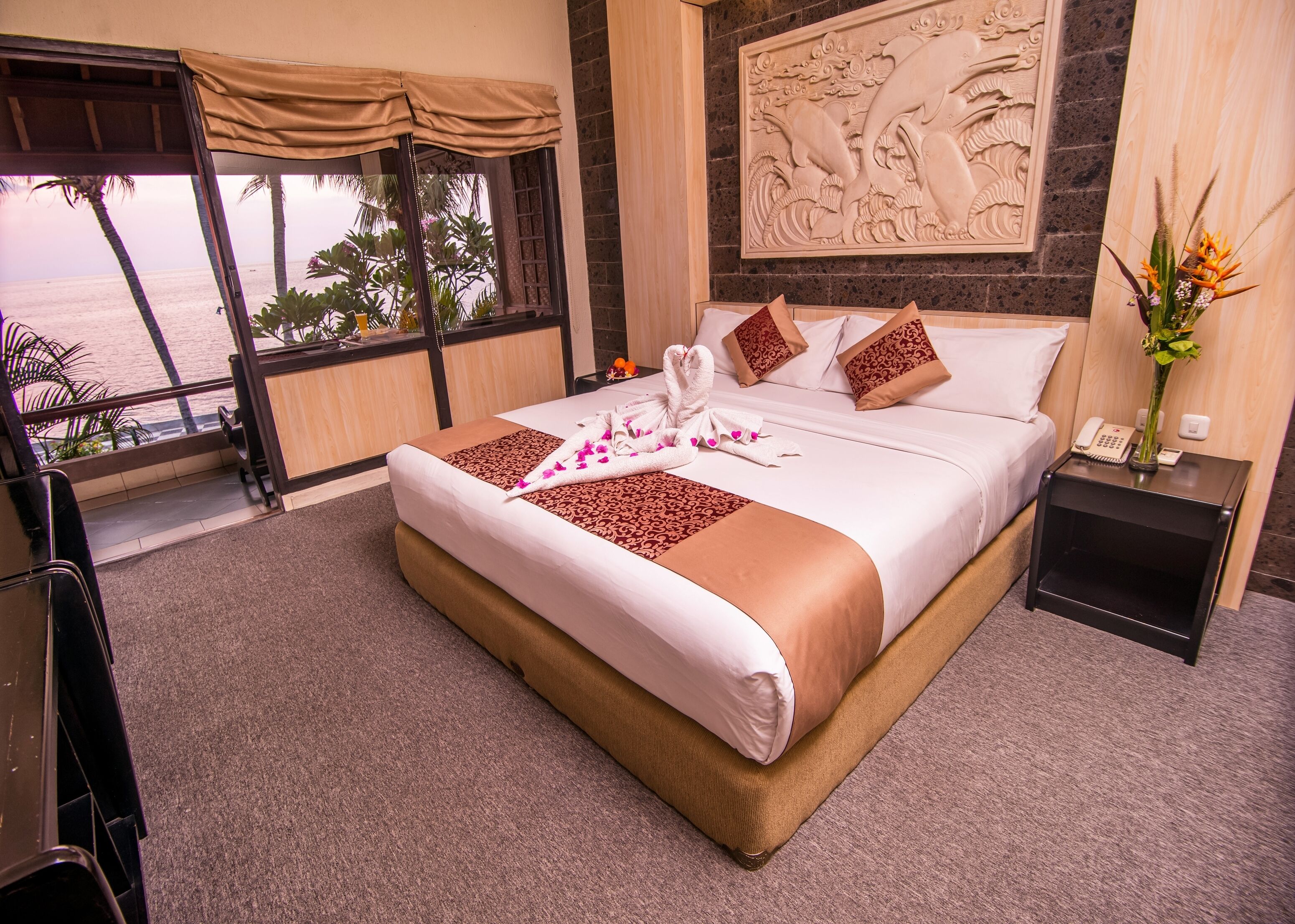 A hotel room with a large bed, a view of the sea, a decorative wall carving, and a small table with a vase of flowers.