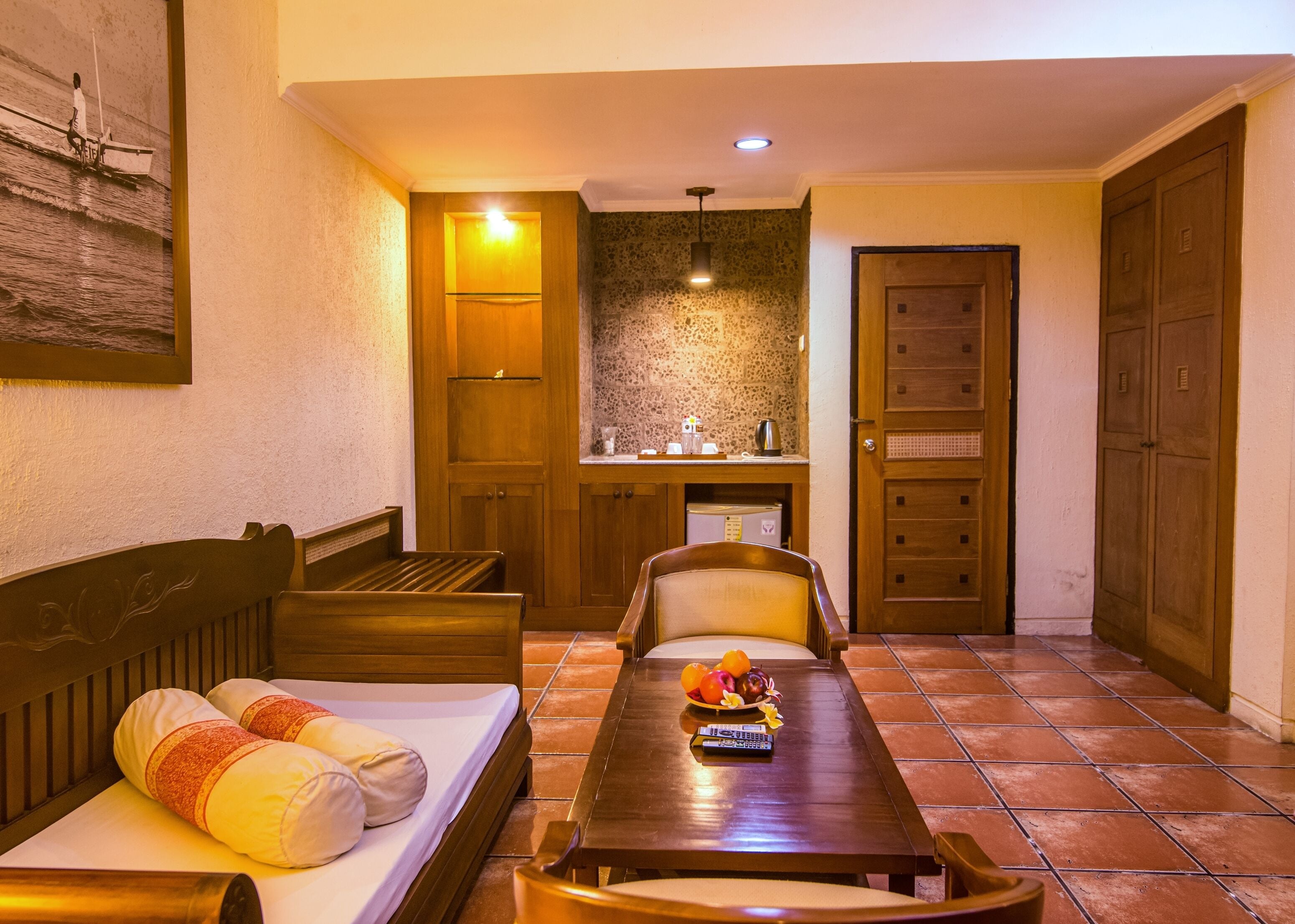 A hotel room with a bed, a wooden table with fruit, a chair, a small kitchen area, and a door.