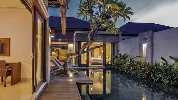 Une maison moderne dotée d’une terrasse en bois qui mène à une piscine à débordement entièrement vitrée, le tout entouré d’une végétation luxuriante.
