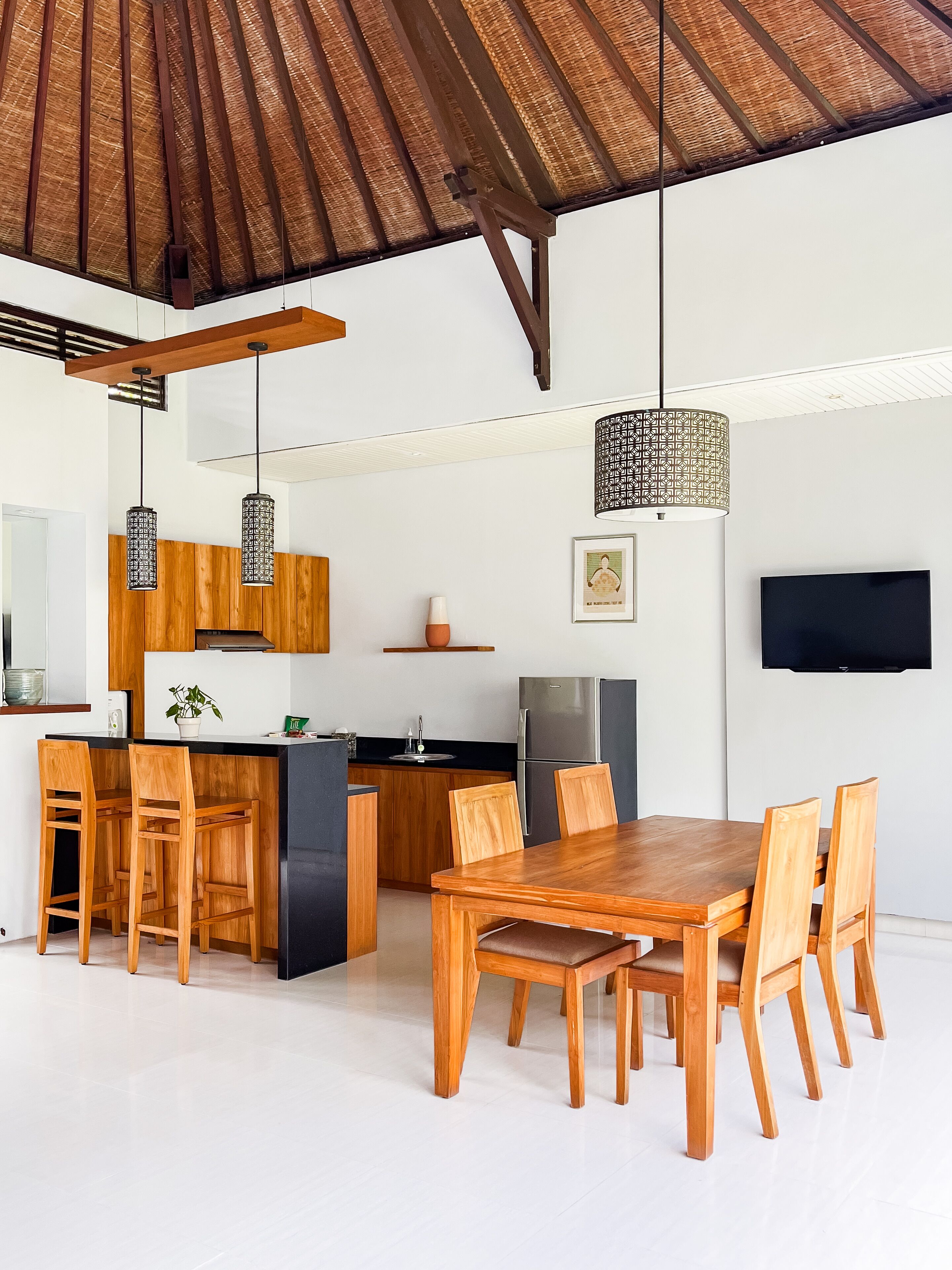 Una cocina moderna con armarios de madera, horno empotrado y un refrigerador grande. Hay un comedor con mesa y sillas de madera, y un televisor montado en la pared.