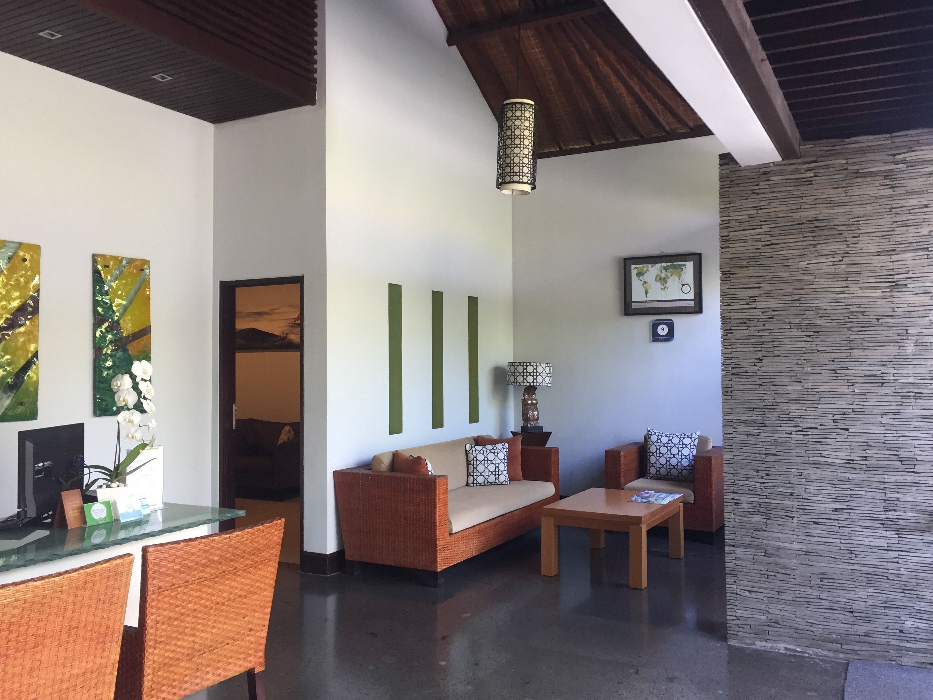 A modern living area with a sofa, two armchairs, a coffee table, and a dining table with wicker chairs. There are abstract paintings on the walls, a decorative lamp, and a stone accent wall.