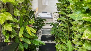 Un étroit sentier mène à une salle de bain dotée d’une baignoire, le tout étant entouré d’une végétation luxuriante.